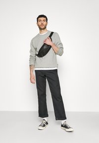 Felpa grigia, borsa a tracolla nera, pantaloni scuri, sneakers nere, calzini bianchi. Modello in piedi con uno sfondo neutro e un leggero sorriso.