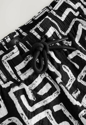 Melnbaltas peldkostīms, kas izgatavots no viegla auduma un ar zigzaga rakstu. Iekļauta elastīga josta ar melnu auklu.