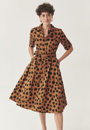 Donna con capelli ricci e corti, che indossa un vestito marrone al ginocchio con grandi pois neri, maniche corte, bottoni e una cintura in vita, in piedi con le mani nelle tasche.
