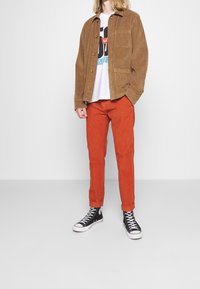 Veste en velours côtelé marron, pantalon orange, t-shirt graphique blanc en dessous. Baskets montantes noires avec semelle blanche. Design simple, coupe décontractée.