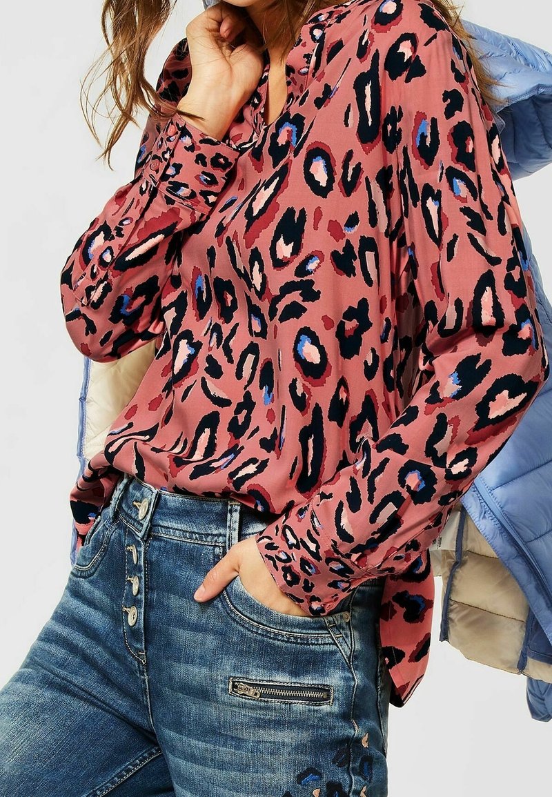 Blouse à imprimé léopard rose avec des manches longues, des poignets à boutons et une coupe décontractée. Portée avec un jean en denim bleu comportant une poche zippée.