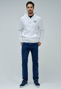 Sudadera blanca con capucha y texto "SALSA JEANS", con bolsillo tipo canguro, combinada con jeans oscuros y zapatillas azul marino. Fondo liso.