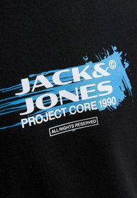 Crna tkanina s bijelim tekstom "JACK & JONES PROJECT CORE 1990" na pozadini plave mrlje boje s natpisom "SVA PRAVA ZADRŽANA" ispod.