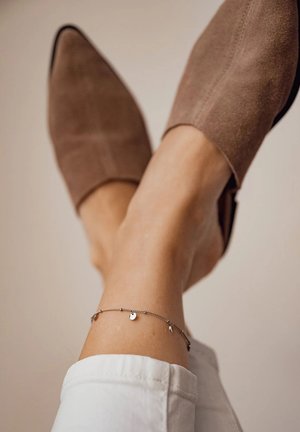 Zapatos de ante marrón sin cordones combinados con una delicada pulsera de tobillo plateada que presenta pequeños encantos, sobre un fondo de color claro.