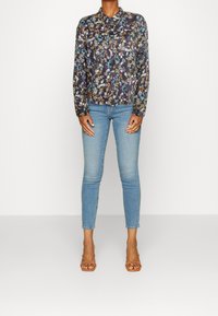 Blommig långärmad blus i mörkt tyg med krage, tillsammans med åtsittande ljusblå jeans. Modellen bär tanfärgade sandaletter med remmar.