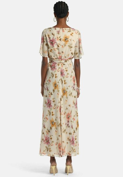 Hölgy fonott hajjal, krém színű virágos maxi ruhában, fodros ujjal és bézs magas sarkú cipővel, háttal állva.
