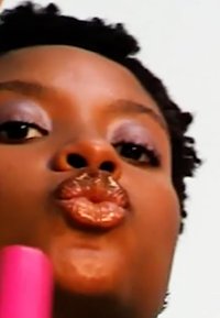 Gros plan sur une personne avec des cheveux bouclés, portant un fard à paupières mauve scintillant et un rouge à lèvres brun brillant, tenant un objet rose devant elle.