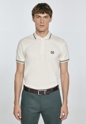 Kremo spalvos polo marškiniai su kontrastinga juoda apykakle ir rankovių juostelėmis. Yra siuvinėtas logotipas ant krūtinės. Dėvimi su žaliais kelnais.