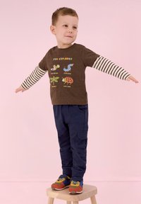 Brun bomulls-T-shirt med färgglada insekt-grafik och text, parat med marinblå cargobyxor och flerfärgade sportskor.