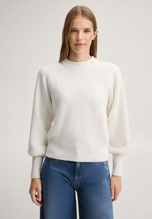 Vrouw met steil lichtbruin haar draagt een witte gebreide trui met ballonmouwen en blauwe jeans, staand tegen een effen lichte achtergrond.