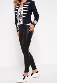 Granatowa kurtka w stylu wojskowym z białymi paskami i guzikami, w zestawie z czarnymi legginsami ze sztucznej skóry oraz białymi butami wsuwanymi z czarnymi akcentami.