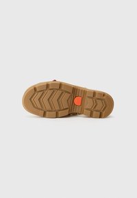 Bruine rubberen zool van een schoen met diepe loopstructuren en een klein oranje cirkelvormig logo nabij het booggebied op een witte achtergrond.