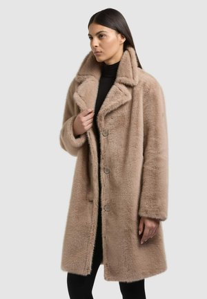 Femme aux longs cheveux foncés portant un manteau en fausse fourrure marron clair longueur genou sur un top noir, sur un fond clair uni.