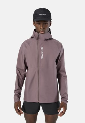 Mężczyzna w różowofioletowej kurtce z kapturem z pionowym logo i czarnej czapce, stojący przodem na jednolitym tle.