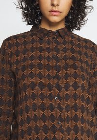 Button-up shirt in black and rust with a geometric fan pattern. Features a pointed collar and front buttons. Made from a textured fabric.
