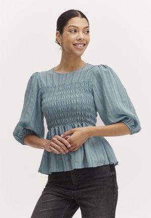 Lichtblauwe blouse met een geweven smock-borst, pofmouwen en een uitlopende zoom. Gemaakt van lichte stof met verticale strepen.