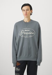 Grå crewneck sweatshirt med hvit "IRON Paradise" tekst. Myk stoff, ribbestrikkede mansjetter og halslinning, avslappet passform. Kombinert med mørke bukser med lapper.