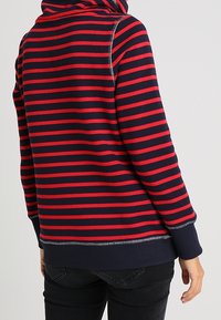 Pullover a righe rosse e blu con cappuccio, caratterizzato da un orlo e polsini a costina. Il tessuto sembra morbido, con dettagli di cucitura visibili.