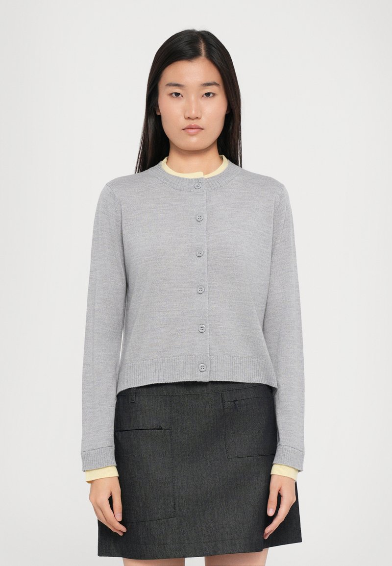 Cardigan grigio a maglia con scollatura rotonda, chiusura frontale con sei bottoni e polsini a costine, sopra uno strato interno giallo, abbinato a una gonna scura.