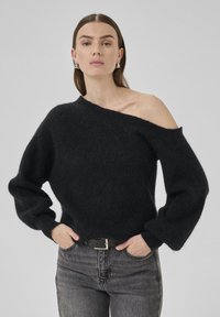 Sort sweater i blødt, fuzzy materiale med off-shoulder design og lange, voluminøse ærmer. Parret med mørke jeans og et sort bælte.