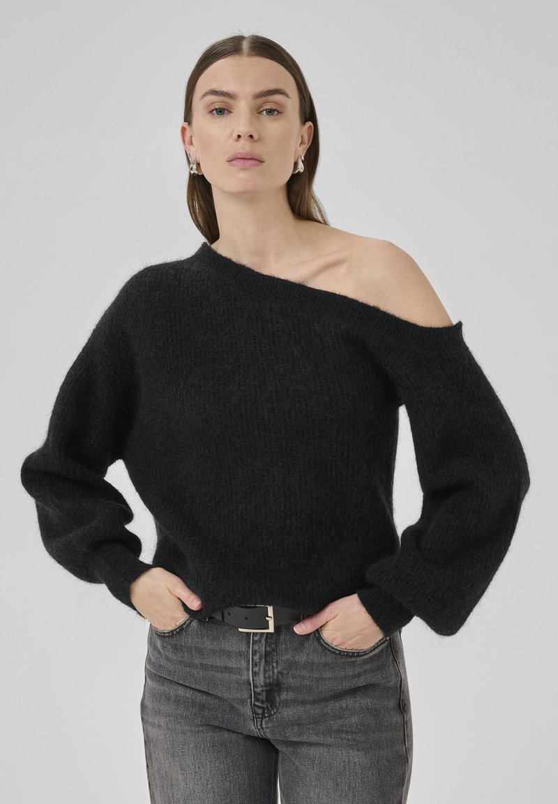 Sort sweater i blødt, fuzzy materiale med off-shoulder design og lange, voluminøse ærmer. Parret med mørke jeans og et sort bælte.