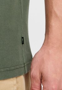 Camiseta verde oliva con un diseño ajustado, hecha de tela suave. Presenta una pequeña etiqueta negra en el lado izquierdo, cerca del dobladillo. Una mano es visible al lado.