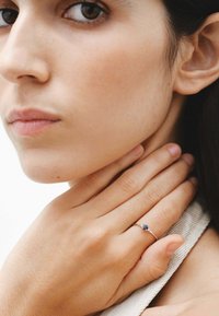 Anillo de plata con una piedra azul, acentuado con piedras claras más pequenas. La mano del modelo descansa sobre su cuello contra un fondo neutro.