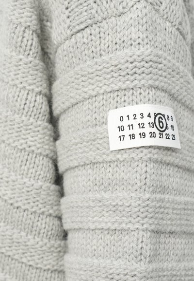 Texturerad, ljusgrå stickad tröja med horisontell ribbad struktur. Har en etikett med storlek och siffror i svart på vit bakgrund.