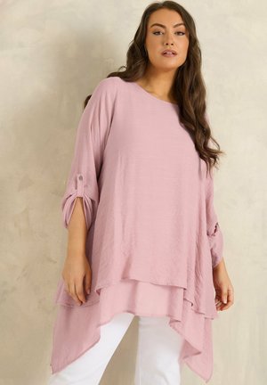 Donna che indossa una tunica asimmetrica rosa chiaro leggera con maniche a 3/4 e pantaloni bianchi, su uno sfondo beige uniforme.