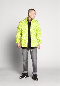Neon gul leopardmønster jakke, glidelås foran, elastiske mansjetter, lett materiale, kombinert med en svart skjorte og gråjeans, svarte sko.