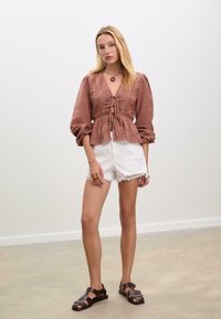 Rostfärgad, texturerad blus med puffärmar och dragsnörning i midjan, i kombination med vita slitna shorts och mörkbruna sandaler.