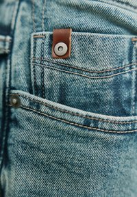 Kieszeń w dżinsach wykonana z denimu z brązową skórzaną naszywką z metalowym zatrzaskiem. Jasnoniebieski, teksturowany materiał z widocznymi przeszyciami i śladami zużycia.