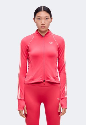 Frau trägt eine enganliegende rosa Jacke mit Reißverschluss und passende Leggings mit weißen Streifen und Adidas-Logos vor einfachem Hintergrund.