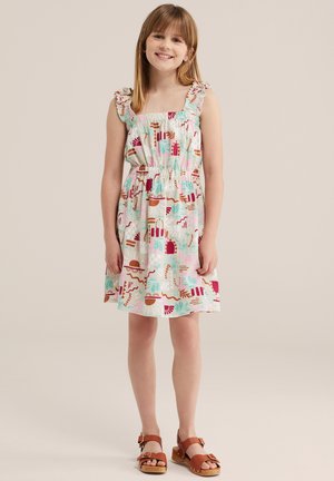 Ragazza sorridente con capelli lisci castano chiaro indossa un vestito bianco senza maniche con motivi astratti colorati e sandali marroni, in piedi su uno sfondo beige.