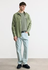 Νεαρός άνδρας που φοράει πράσινο καρό πουκάμισο πάνω από πράσινο πλεκτό μπλουζάκι, ανοιχτόχρωμο τζιν, μαύρα παπούτσια, στέκεται με το ένα χέρι στην τσέπη σε απλό φόντο.