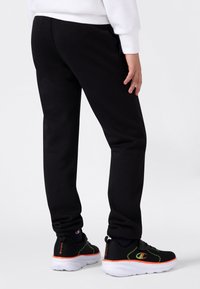 Kind, das schwarze Sweatpants und schwarze Sneaker mit neongrünen und orangefarbenen Akzenten trägt, steht auf einem weißen Boden vor einem weißen Hintergrund.