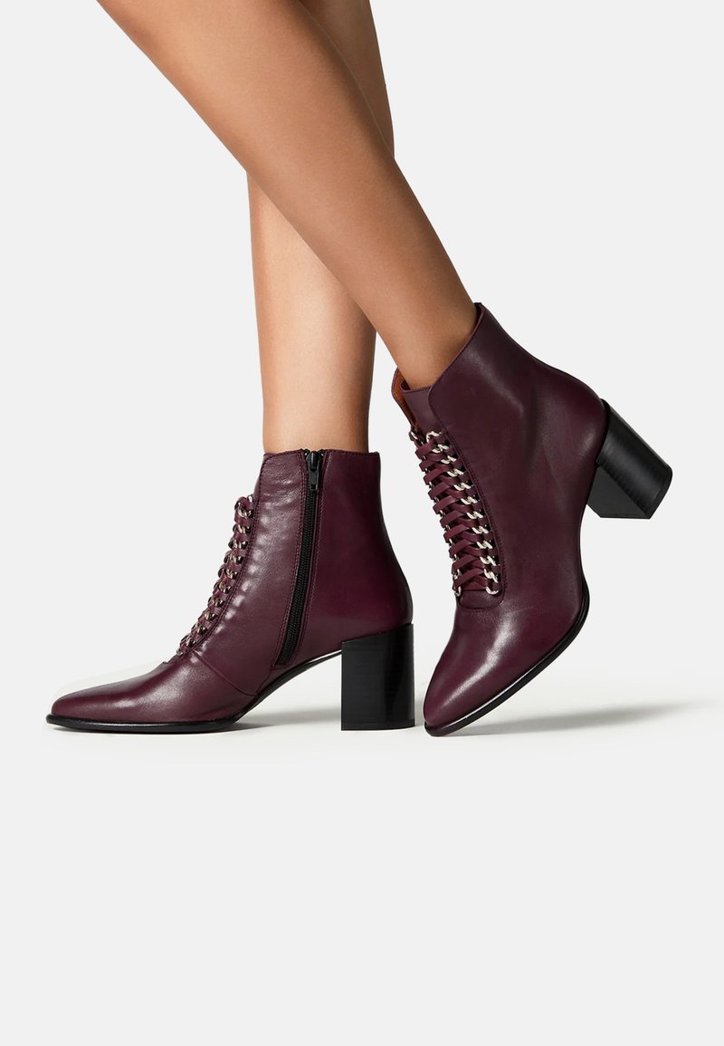 Picioare purtând cizme de piele mov închis, cu fermoar lateral, detalii cu șireturi în față și tocuri pătrate, pe un fundal alb.