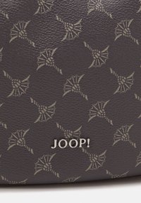 Dunkelgraues strukturiertes Material mit einem sich wiederholenden floralen Muster in einem helleren Farbton. Silbernes Emblem mit der Aufschrift "JOOP!" am unteren Rand.