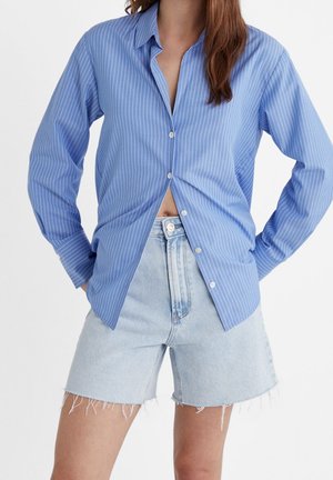 Personne portant une chemise à manches longues à fines rayures bleues partiellement boutonnée sur un short en jean léger coupé, sur un fond uni.