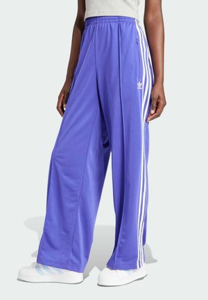 Personne portant un pantalon de survêtement Adidas violet avec des bandes blanches, un haut gris et des baskets blanches, debout sur un fond clair.
