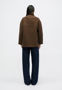 Καφέ σακάκι από μείγμα μαλλιού με ψηλό γιακά, χαλαρή γραμμή και μανίκια μέχρι τον αγκώνα. Συνδυασμένο με φαρδιά τζιν παντελόνια σκούρου χρώματος.