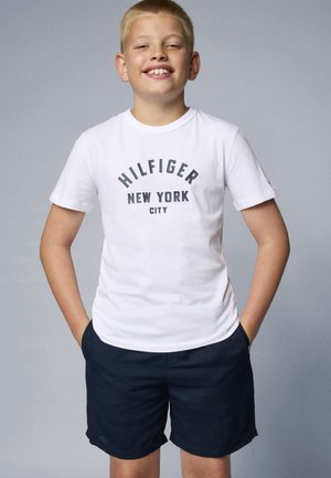 Fehér pamut póló, sötétszürke "HILFIGER NEW YORK CITY" felirattal, tengerészkék sorttal párosítva. A modell keze a zsebében áll.
