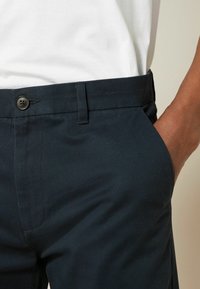 Marineblaue Hose aus strukturiertem Stoff, mit Knopfverschluss, Seitentaschen und schmalem Schnitt.