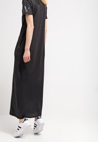 Vestido maxi negro con mangas cortas, tejido suave, corte relajado y una abertura hasta la rodilla. Combinado con zapatillas blancas con rayas negras.