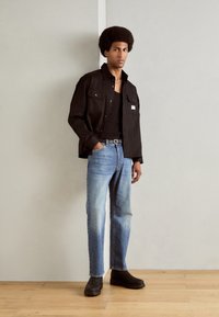 Chemise noire boutonnée sur un débardeur noir, associée à un jean bleu clair et des chaussures noires à enfiler. Textures lisses et coupe décontractée.