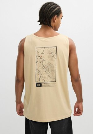 Hombre con el cabello trenzado que lleva una camisa sin mangas beige con un mapa contorneado negro y marcadores rojos etiquetados como "White Peak" en la parte trasera.