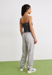 Un pantalon de survêtement gris associé à un débardeur smocké noir à fines bretelles. L'arrière-plan est un mur uni avec un sol vert.