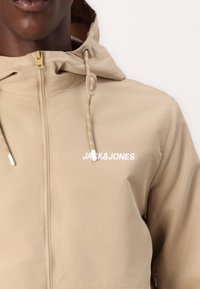Beige huva jacka med framdragkedja, dragsnodd-accents och en synlig "JACK&JONES" logotyp i vitt. Slät tygstruktur.