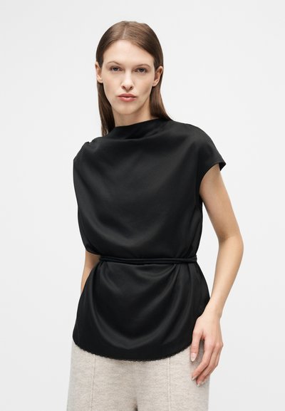 Γυναίκα με ίσια καστανά μαλλιά φοράει φαρδιά μαύρη μπλούζα με σφηκοφωλιά στη μέση και μπεζ υφασμάτινο παντελόνι, στέκεται μπροστά σε απλό φωτεινό φόντο.