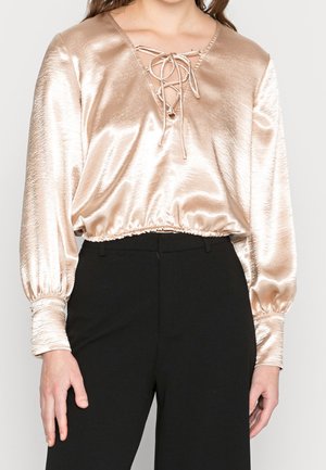 Vrouw draagt een glanzende champagneton satijnen blouse met vetersluiting bij de halslijn en lange pofmouwen, gecombineerd met zwarte broek.